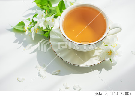 Cup of hot tea with white jasmine flowers and green leaves on a white surface with soft shadows Cup of hot tea with white jasmine flowers and green leaves on a white surface with soft shadows 143172805