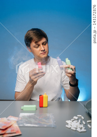 Drug dealer holding two colorful electronic cigarettes surrounded by smoke with more vapes, pills, money and plastic baggies on table against isolated blue background Drug dealer holding two colorful electronic cigarettes surrounded by smoke with more vapes, pills, money and plastic baggies on table against isolated blue background 143172876