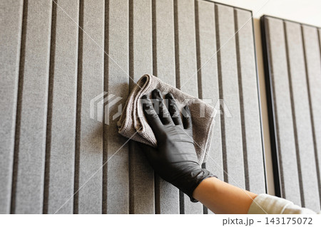 Person wearing black protective glove cleaning a textured grey acoustic wall panel with a cloth 143175072