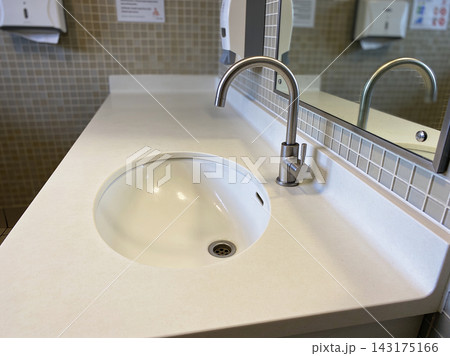 Modern stainless steel faucet and white ceramic sink in a clean public restroom vanity area Modern stainless steel faucet and white ceramic sink in a clean public restroom vanity area 143175166