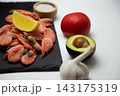 There is a slice of lemon on a black slate with shrimps, and a blurred salt shaker, tomato, avocado, and garlic are placed next to it on a white background. Concept: deep composition, an emphasis on 143175319