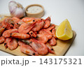A close-up side view of frozen shrimps on a wooden board, two lemon slices diagonally, a salt shaker and a head of garlic next to it. Concept: an expanded set of seafood seasonings. High quality photo 143175321