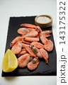 Frozen shrimps in frost, a slice of lemon, and a wooden salt shaker are lying on a black stone board with uneven edges, and a wooden salt shaker is blurred in the background. Concept: cold gastronomic 143175322