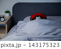 There is a black alarm clock on a wooden bedside table, and in the background a brown bear in a red mask sleeps wrapped in a white blanket under a gray headboard. Concept: the toy aesthetics of sleep 143175323