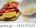 On a white background, there is a bowl of rice, a wooden board with frozen shrimp, a slice of lemon, and a few grains of cereal spilled onto the countertop. High quality photo 143175325