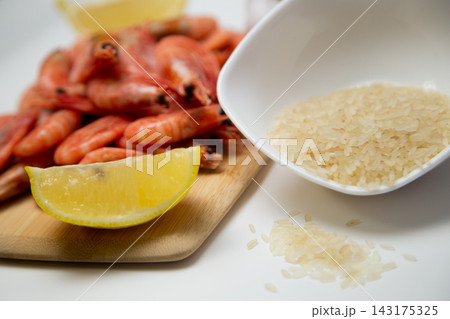 On a white background, there is a bowl of rice, a wooden board with frozen shrimp, a slice of lemon, and a few grains of cereal spilled onto the countertop. High quality photo 143175325