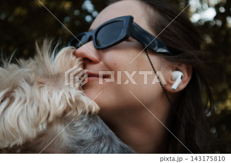 Woman wearing sunglasses and earbuds holds a small dog outdoors, sharing an affectionate moment in warm natural light. Pet companionship. 143175810