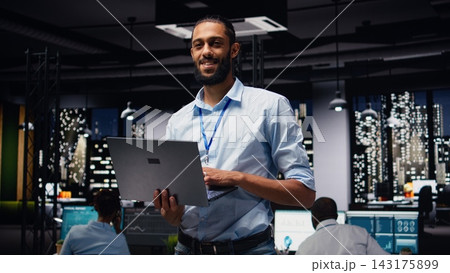 Portrait of smiling middle eastern employee standing in office reading emails on laptop, responding to inbox messages and solving tasks. Man staff analyzing KPIs, interpreting statistical data. 143175899