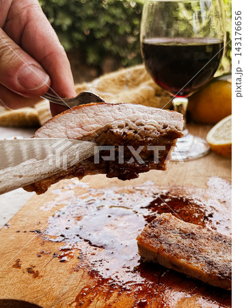 Person carving juicy grilled beef from an Argentine asado with a fork and knife, preparing a delicious gourmet meal with red wine and fresh lemons 143176656