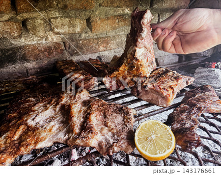 Hand turning sizzling beef ribs and various cuts of asado on a hot charcoal grill, creating a traditional Argentinian barbecue experience with a fresh lemon Hand turning sizzling beef ribs and various cuts of asado on a hot charcoal grill, creating a traditional Argentinian barbecue experience with a fresh lemon 143176661