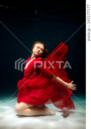 Scarlet clad ballerina gracefully poised beneath water surface Scarlet clad ballerina gracefully poised beneath water surface 143177177