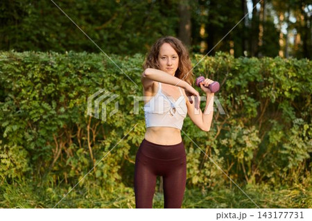 Athletic woman performs shoulder exercises in sunny outdoor setting Athletic woman performs shoulder exercises in sunny outdoor setting 143177731