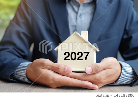 Businessman hands holding wooden House model with 2027 New Year text. Property insurance and real estate concepts Businessman hands holding wooden House model with 2027 New Year text. Property insurance and real estate concepts 143185800