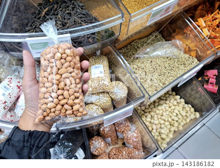 Customer picking packaged raw peanuts in transparent plastic bag. Supermarket rack filled with various spices and grains in clear bins. 143186182