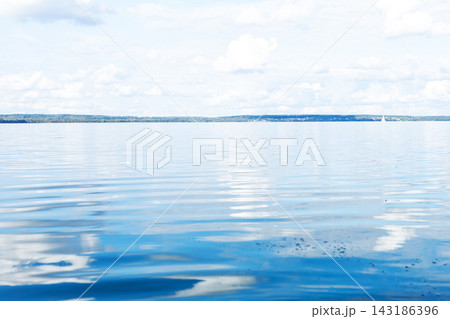 Calm freshwater lake surface reflecting pale blue sky and scattered clouds with distant shoreline at soft daylight serene horizontal waterscape for travel and wellness background Calm freshwater lake surface reflecting pale blue sky and scattered clouds with distant shoreline at soft daylight serene horizontal waterscape for travel and wellness background 143186396