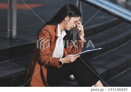 Asian business woman sitting alone on concrete steps, suffering from severe headache, mental stress and deep sadness, contemplating failure after losing her job 143186685