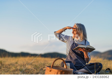 Older woman enjoying peaceful solo picnic and reading in countryside. 143188062