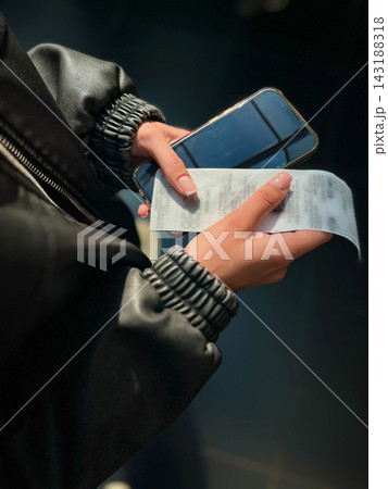 Unrecognizable teenager holding paper receipt and smartphone after making a purchase in mall. Concept of consumer spending, financial transaction tracking, and modern shopping habits. Unrecognizable teenager holding paper receipt and smartphone after making a purchase in mall. Concept of consumer spending, financial transaction tracking, and modern shopping habits. 143188318
