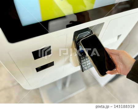 Close up of teenage girl paying for food using mobile phone contactless payment at self service kiosk. Concept of cashless transactions, modern NFC technology, and digital everyday life. Close up of teenage girl paying for food using mobile phone contactless payment at self service kiosk. Concept of cashless transactions, modern NFC technology, and digital everyday life. 143188319