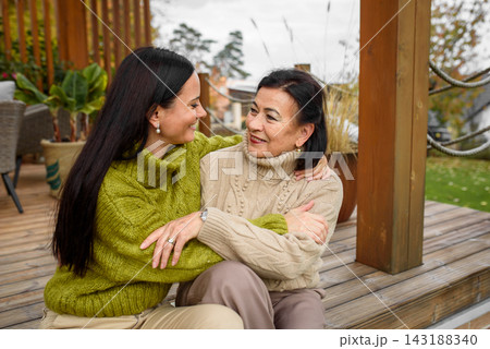 Mature daughter with older mom sitting on wooden patio, hugging. Mother's day concept. Mature daughter with older mom sitting on wooden patio, hugging. Mother's day concept. 143188340