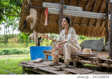 Asian woman farmer resting under thatched shelter holding fan and gloves. 143188659