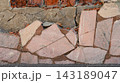 A close-up of a crumbling weathered stone wall with broken light pink granite tiles, cracked concrete, and exposed red bricks. 143189047