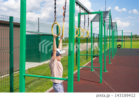 A child is actively playing on gymnastic rings in a vibrant outdoor playground setting, featuring a green metal frame and a wooden fence. The scene is set under a cloudy sky, creating a fun. 143189412