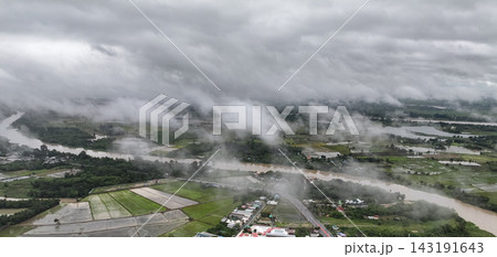 Rain clouds blanket swollen river winding through lush agricultural fields, dense mist rolls across the flooded rural landscape in gloomy monsoon day. Rain clouds blanket swollen river winding through lush agricultural fields, dense mist rolls across the flooded rural landscape in gloomy monsoon day. 143191643