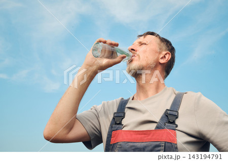 Man in overalls hydrating during work break with blue sky background 143194791