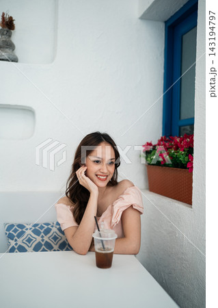 Smiling woman in soft pink off shoulder top sitting at white cafe table with iced drink, relaxed pose beside bright blue window, white wall, patterned cushion, and red flowers Smiling woman in soft pink off shoulder top sitting at white cafe table with iced drink, relaxed pose beside bright blue window, white wall, patterned cushion, and red flowers 143194797