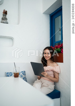 Smiling young woman using tablet while seated on blue bench at bright white cafe terrace beside blue window, pink flowers, and blurred iced drink on table 143194808