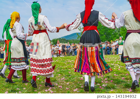 Folk dancers holding hands over rose petals. Women dancers in embroidered dresses hold hands while moving across grass scattered with pink rose petals. Colorful skirts, headscarves, open field Folk dancers holding hands over rose petals. Women dancers in embroidered dresses hold hands while moving across grass scattered with pink rose petals. Colorful skirts, headscarves, open field 143194932