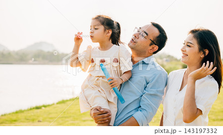 Asian family having fun playing together on spring meadow field grass in park, Happy parents father carrying daughter and a child blowing soap bubbles enjoying outdoors together 143197332