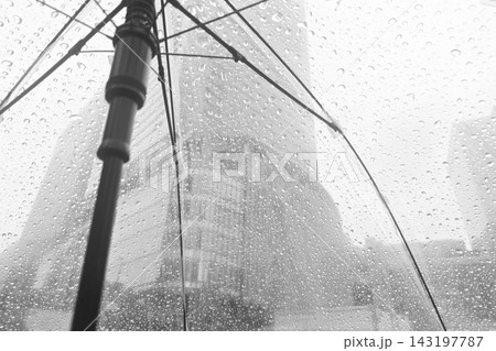 Close-up view of raindrops on a transparent umbrella with a blurred modern city building background on a rainy day. Close-up view of raindrops on a transparent umbrella with a blurred modern city building background on a rainy day. 143197787