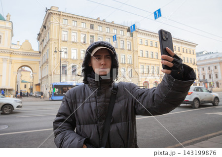 Young man taking selfie with smartphone on city street in winter. Urban tourist photography and social media lifestyle concept. Travel blogger creating content outdoors Young man taking selfie with smartphone on city street in winter. Urban tourist photography and social media lifestyle concept. Travel blogger creating content outdoors 143199876