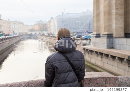Student man standing on bridge and looking at city canal. Young guy wearing winter jacket and cap enjoys urban view. Travel, leisure and reflection Student man standing on bridge and looking at city canal. Young guy wearing winter jacket and cap enjoys urban view. Travel, leisure and reflection 143199877