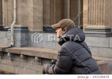 Young man in cap and winter jacket sitting near historic building with columns. Thoughtful guy resting on city street. Urban lifestyle and cold season concept. Young man in cap and winter jacket sitting near historic building with columns. Thoughtful guy resting on city street. Urban lifestyle and cold season concept. 143199878