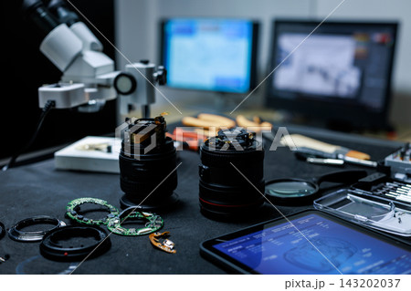 Camera lenses, repair tools and small electronic parts on a workbench beside a microscope and computer screens in a professional photography equipment service workshop. 143202037
