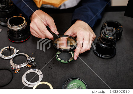 Close-up of a camera technician repairing a disassembled lens on a workbench, using a magnifying glass to inspect small electronic parts. Close-up of a camera technician repairing a disassembled lens on a workbench, using a magnifying glass to inspect small electronic parts. 143202059