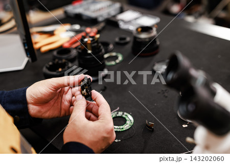 Close-up of hands repairing a small camera lens part on a workbench with tools, lens elements and equipment in a blurred workshop. Close-up of hands repairing a small camera lens part on a workbench with tools, lens elements and equipment in a blurred workshop. 143202060