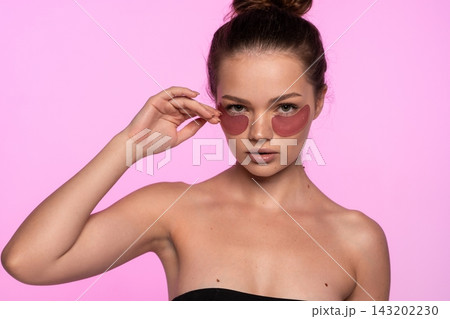Young woman applying pink hydrating eye patches for a cosmetic skin treatment, promoting beauty and self-care Young woman applying pink hydrating eye patches for a cosmetic skin treatment, promoting beauty and self-care 143202230