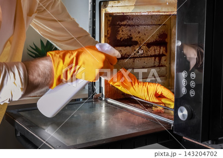 Man in protective gloves cleaning dirty oven with sponge and spray. He performs household chores, doing thorough grease removal and rust cleaning for the kitchen appliance at home. 143204702