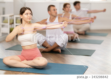 Active young girl wearing sport clothes using resistance band during group workout sitting on mat in gym 143206642