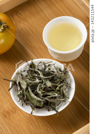 High angle view of Chinese white tea leaves in a white saucer and a cup of freshly brewed tea on a wooden tray 143211544