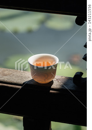 Chinese teacup with dragon relief filled with amber tea on a wooden balcony overlooking a blurred garden background Chinese teacup with dragon relief filled with amber tea on a wooden balcony overlooking a blurred garden background 143211556
