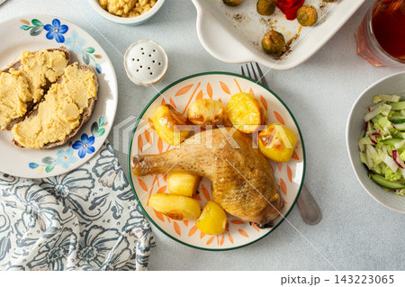 A baked chicken leg rests on a plate with potatoes. Fresh salad, roasted Brussels sprouts, and chickpea bread are arranged on the table. It looks like a wholesome meal 143223065