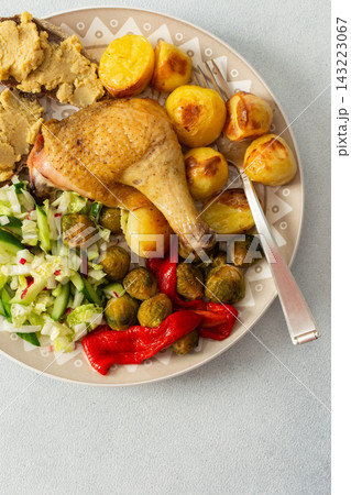 A plate holds baked chicken leg, potatoes, fresh salad, roasted Brussels sprouts, and chickpea bread. This meal is ready to eat at a dining table 143223067