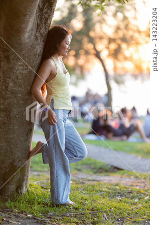 Woman relaxing barefoot leaning against tree at sunset Woman relaxing barefoot leaning against tree at sunset 143223204