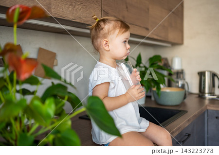 Happy baby girl sitting at the table in the kitchen and drinking milk Happy baby girl sitting at the table in the kitchen and drinking milk 143223778