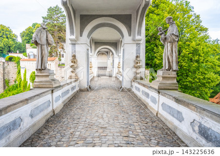 The Coat Bridge connects the Upper Castle and the Baroque Castle Theatre in Cesky Krumlov. Statues line the sides of the stone pathway surrounded by green trees. The Coat Bridge connects the Upper Castle and the Baroque Castle Theatre in Cesky Krumlov. Statues line the sides of the stone pathway surrounded by green trees. 143225636